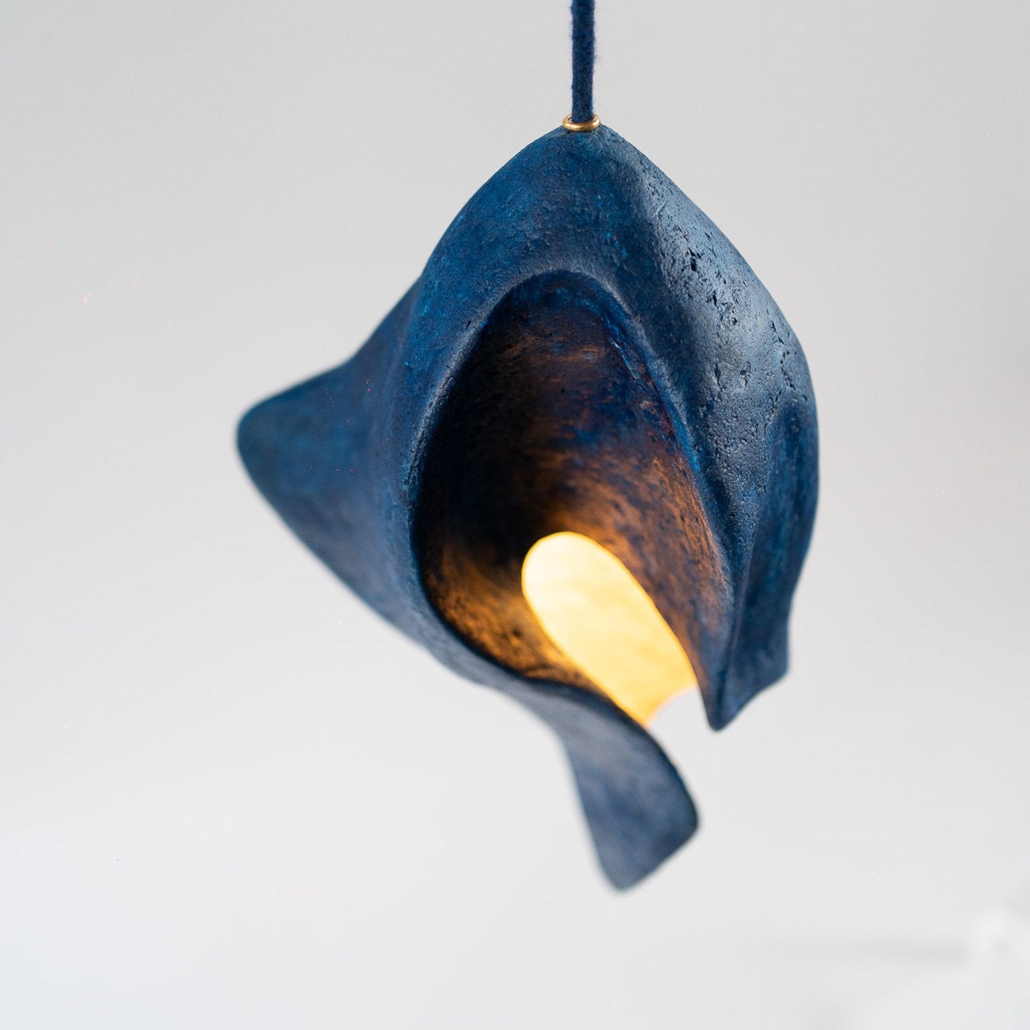 Blue paper mache pendant light with a glowing bulb on a white background that has a sculptural shape and soft stone texture