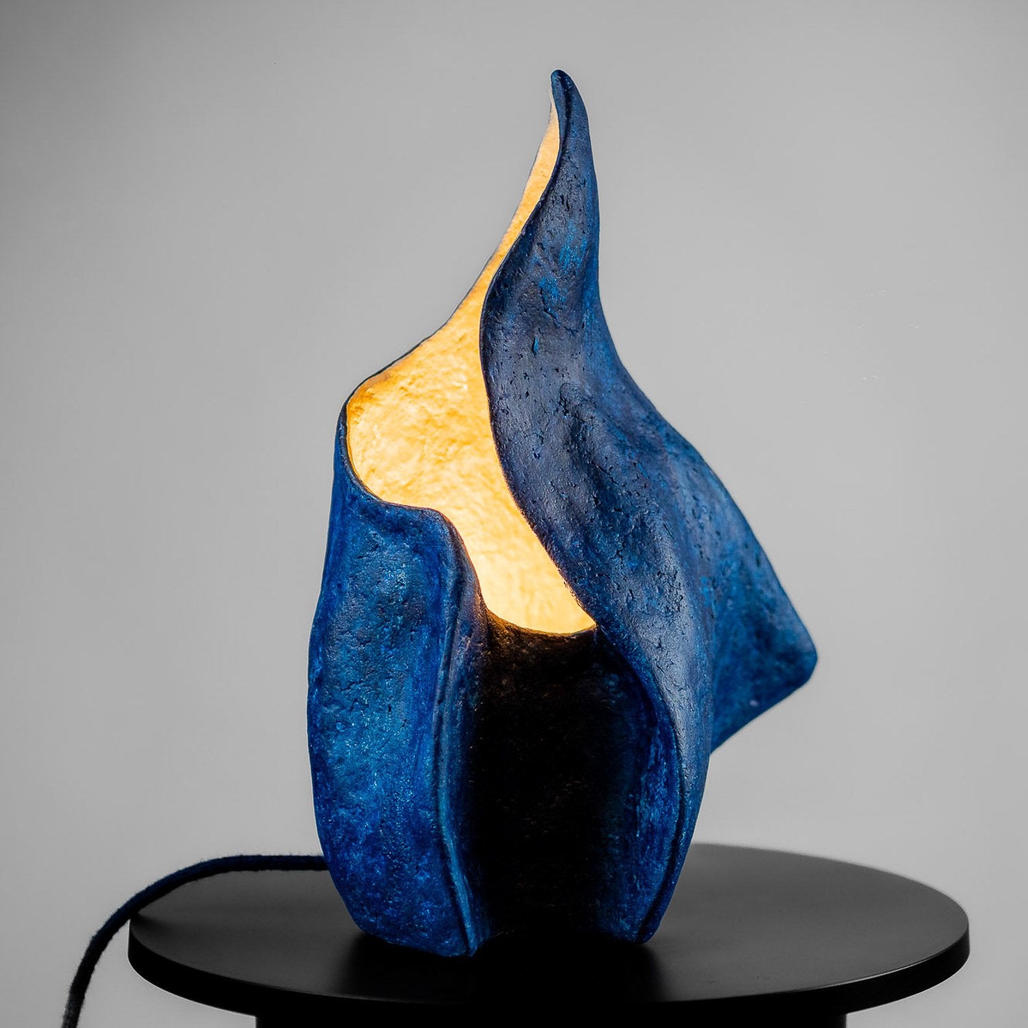 Blue curvy artistic sculpture, a textured table lamp with a warm yellow light on a gray background