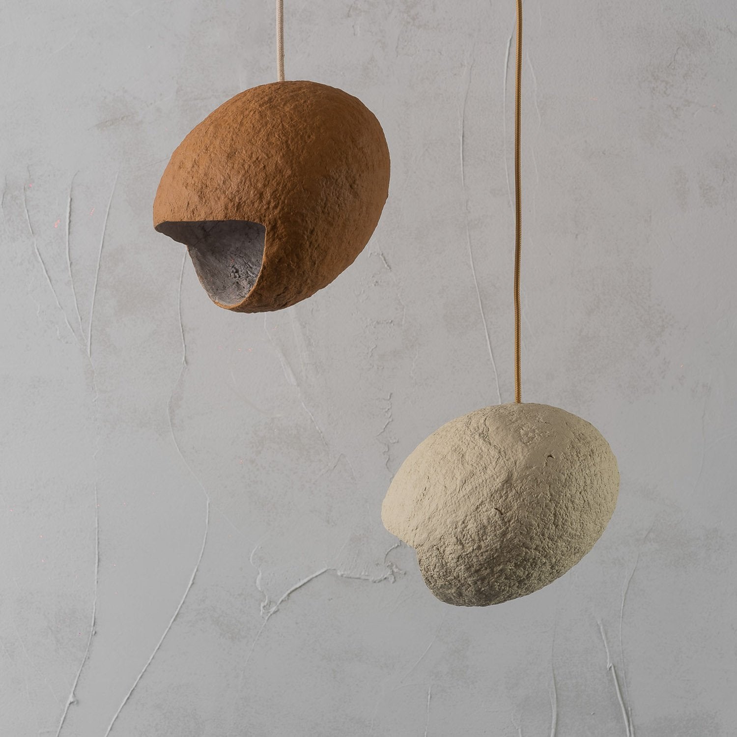Two spherical pendant lights inspired by the nest of birds hanging against a gray textured wall. Lamps are made from paper mache painted with natural pigments of terracotta and beige
