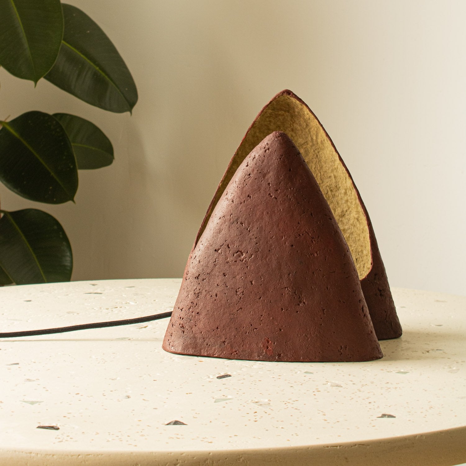 Soft paper mache cone-shaped table lamp on a microcement surface with a plant in the background. Burgundy and beige natural color made by Lavinia Erghelan