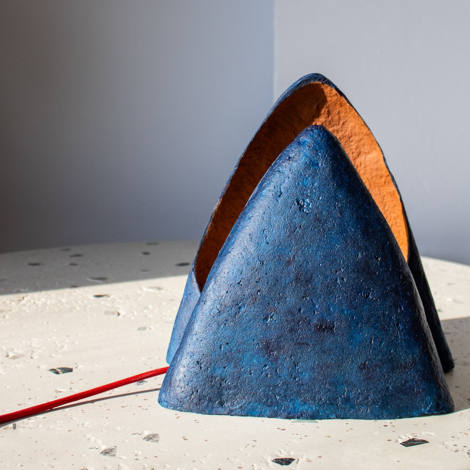 dark blue paper mache with soft texture table lamp, cone-shaped, sculptural triangle looks like mountain. Interior terracotta color and red cord.