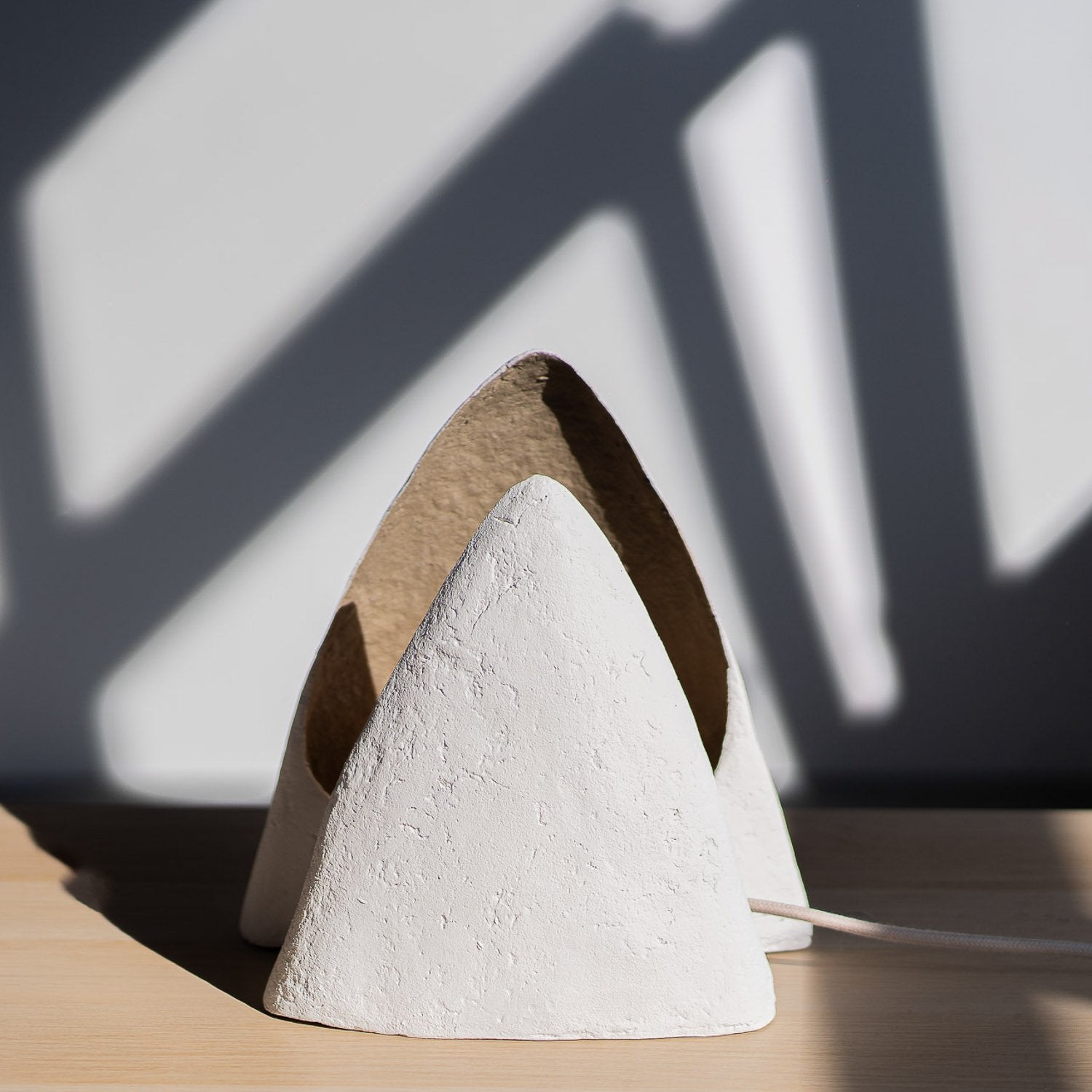 White textured cone-shaped object on a wooden surface with shadows on the wall. Paper mache table lamp with smooth texture hand painted with natural pigments